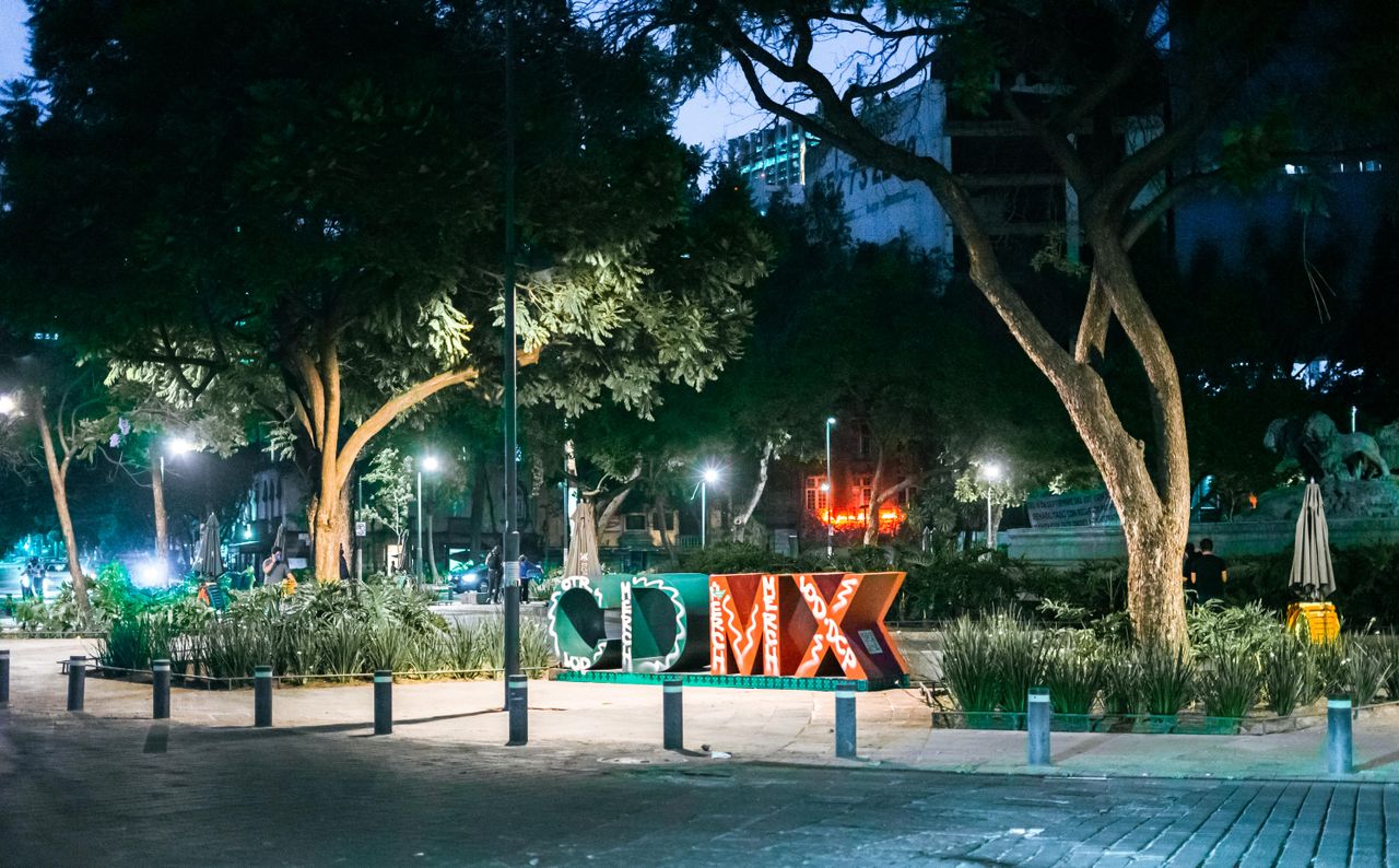 Night scene of the iconic CDMX sign illuminated in a city park in Mexico City, surrounded by trees and ambient lights.