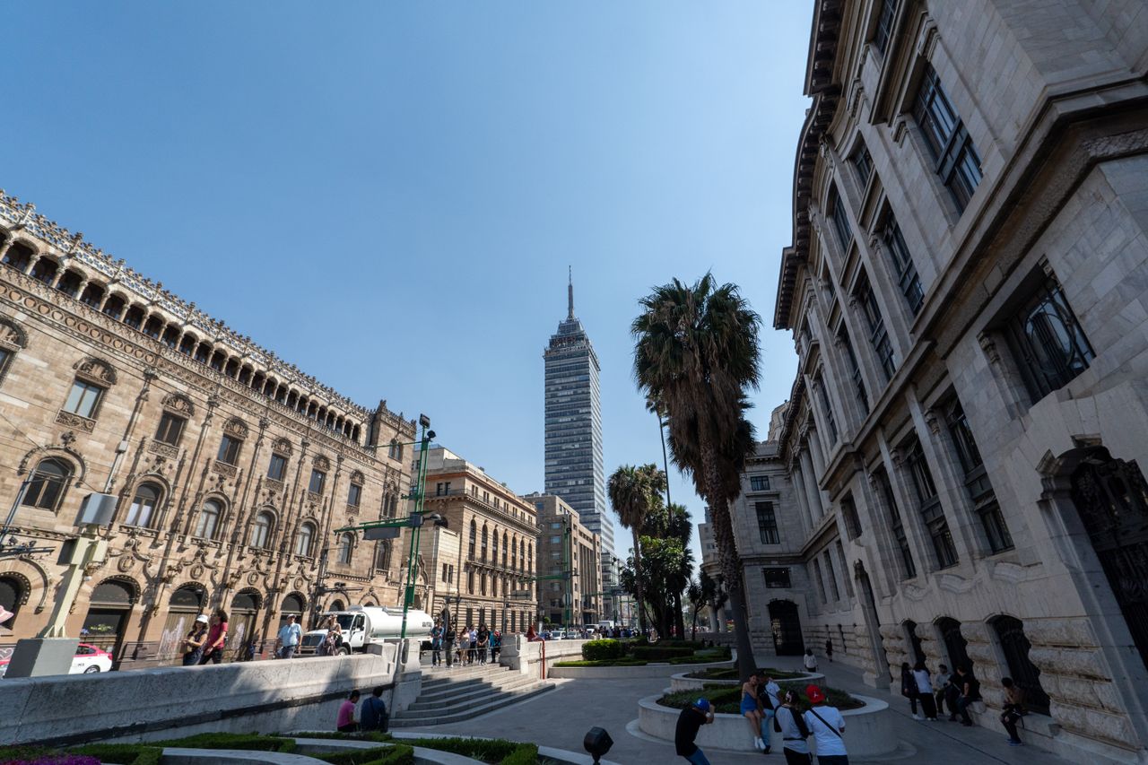 View of Torre Latinoamericana among historic buildings in Mexico City with people and palm trees under a clear sky.